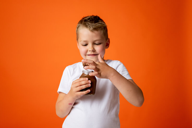 Bambino su sfondo arancione con un vasetto di marmellata in mano, in proconto di assaggiarla con le dita